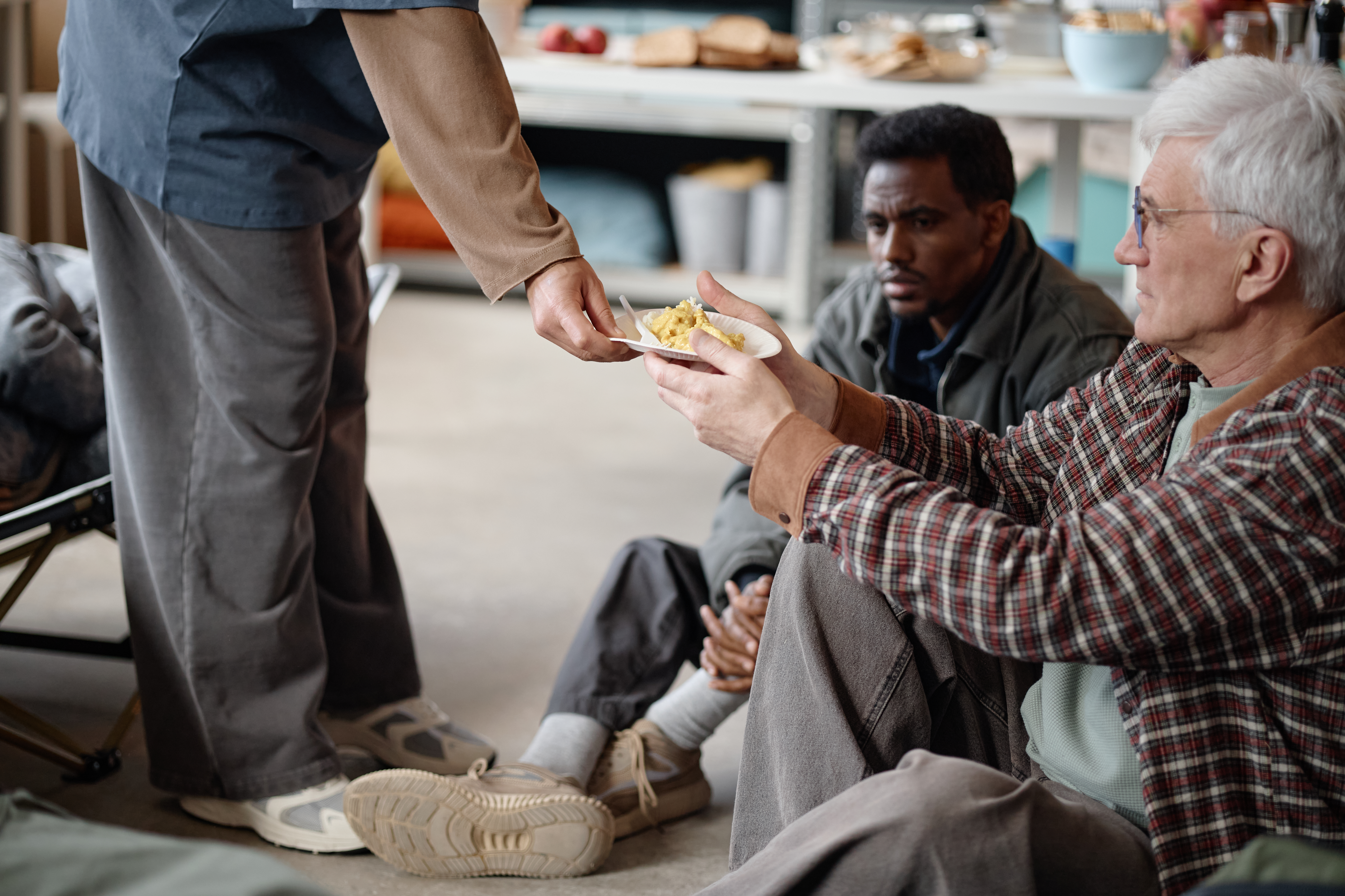 Volunteer handing meal to senior Caucasian man while diverse men sitting in humanitarian aid center, supporting hunger relief and social care. Ideal for charity campaign or nonprofit outreach